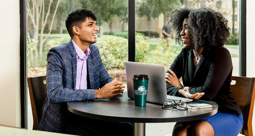 Two people sit at a round table with a laptop, a notebook, and a coffee tumbler, engaged in conversation in a well-lit indoor setting near large windows.